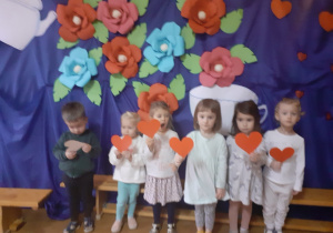 Zdjęcie przedstawia dzieci trzymające czerwone papierowe serca na tle dekoracji z kolorowych kwiatów